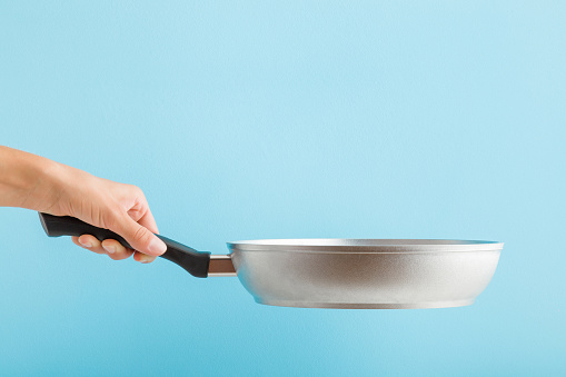 Young adult woman hand holding new aluminium frying pan on light blue background. Pastel color. Closeup. Cooking concept. Side view.