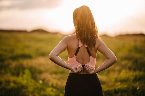 A rear view of a young adult woman adjusting her sports bra and preparing for her morning jog on the hilly meadow