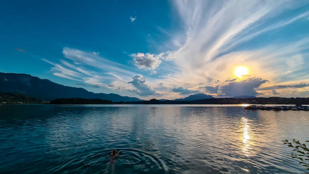 Faakersee - A woman swimming in the Lake Faak in Austria. The lake is surrounded by high Alpine peaks. The sun in slowly setting A woman swimming in the Lake Faak in Austria. The lake is surrounded by high Alpine peaks. The sun in slowly setting behind the mountains. Lots of clouds. Calm surface reflects the sunbeams. Happiness villach stock pictures, royalty-free photos & images