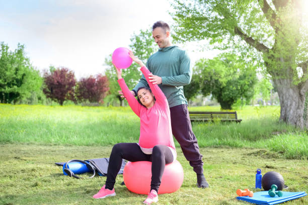 personal trainer sorridente e una donna incinta felice che fa esercizio yoga all'aperto in un parco - la ragazza fa allungamenti muscolari seduti sulla palla da palestra - concetto di allenamento, fitness, in tempo prenatale - yoga ball foto e immagini stock