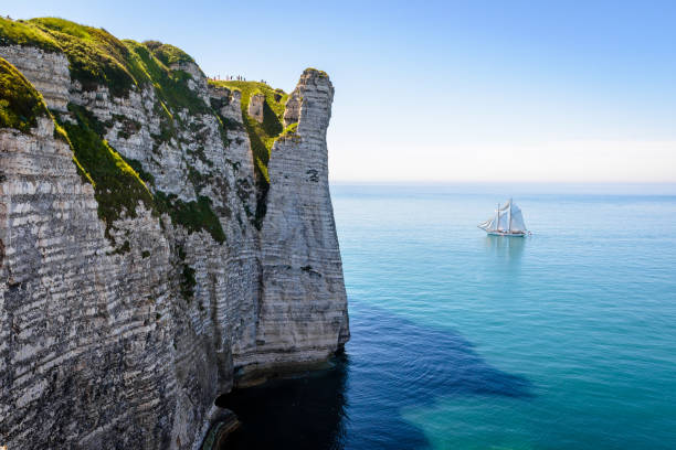 A white sailing ship is cruising, in full sail, off the Aval cliff in Etretat, Normandy, on a calm sea by a sunny day.