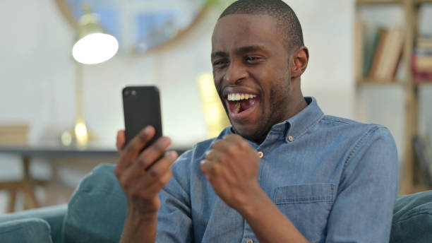 istock Young African Man Celebrating on Smartphone at Home 1329204612