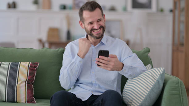 istock Young Man Celebrating Success on Smartphone on Sofa 1329204766