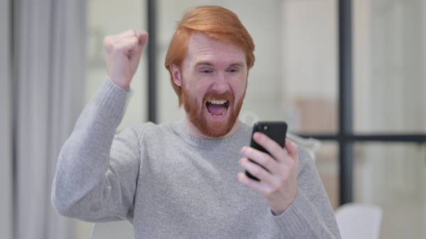 istock Young Beard Redhead Man Celebrating on Smartphone 1329206995