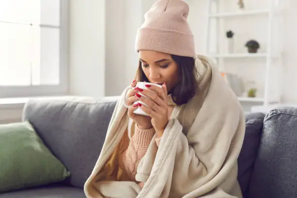 Photo of Woman wearing a knitted sweater and hat sitting on the sofa wrapped in a plaid drinking hot coffee.