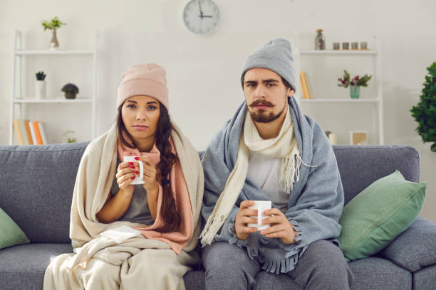 Upset young couple having problem with central heating or suffering from cold or flu Dissatisfied young couple having problem with central heating, sitting on sofa at home, freezing, drinking hot tea trying to warm up. Sick man and woman wrapped in blankets suffering from cold or flu home heating stock pictures, royalty-free photos & images