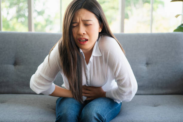 unhappy asian woman sitting on the sofa and holding on stomach suffering. abdominal pain that comes from menstruation, diarrhea, or indigestion. sickness and healthcare concept - colic imagens e fotografias de stock