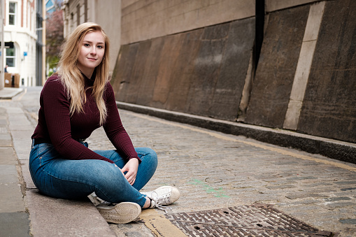 Young woman sitting on the floor and looking to camera in a very narrow street. She is wearing a maroon pullover and blue jeans. The street is stone pavimented.