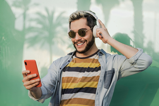 Young hipster man listening music on mobile phone app while wearing headphones outdoor - Focus on face