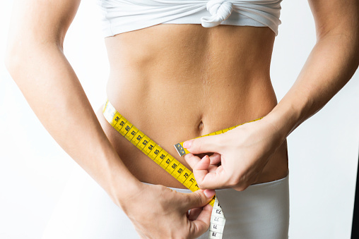 Belly of an unrecognizable slim caucasian woman wearing sportswear is measuring her waist in front of white background. Representing being healthy and slim after regular workout and healthy lifestyle. Note : Image is not body shape retouched.