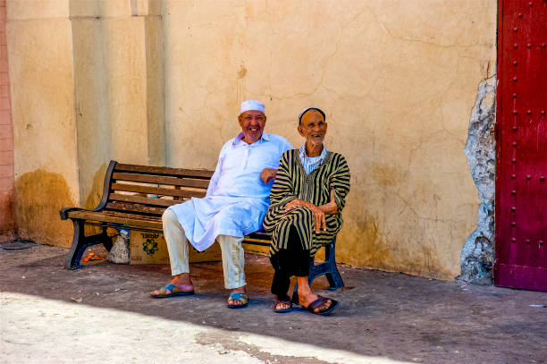 Two men, obviously friends relaxing on a bench along a busy street in \nMarrakech.  Their expressions suggest that they knew many of the passer-by.  These men are seated in the Medina (old city) of Marrakech in the area know as a souk.  A souk is an outdoor market where both tourists and locals can shop for a variety of items.  Many people relax as these men are doing within the medina and chat with friends and neighbors exchanging the days news.