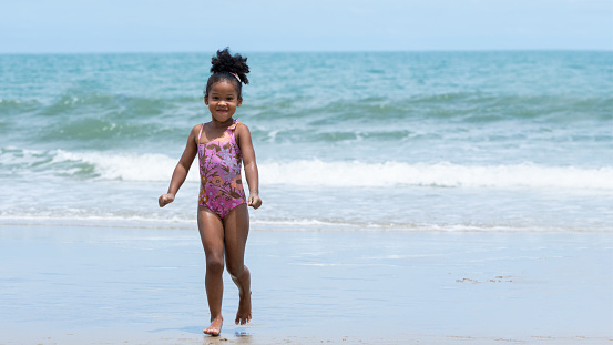 Joyful African American kid running at the sea on the seashore of a tropical island. A little girl  playing fun on vacation at blue sky beach on a sunny day. Summer holiday party.