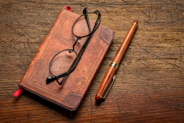 small leather-bound journal with a stylish pen and reading glasses on a rustic wooden table, journaling concept