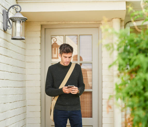 joven de pie fuera de la puerta de su casa y enviando mensajes de texto en su teléfono - home sale fotografías e imágenes de stock