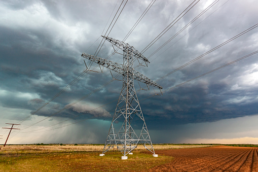 Dangerous severe storm approaches high voltage electricity power lines in agricultural field: steel pylons carrying multiple power cables appear to stand defiant as the maelstrom bears down. Wide angle view
