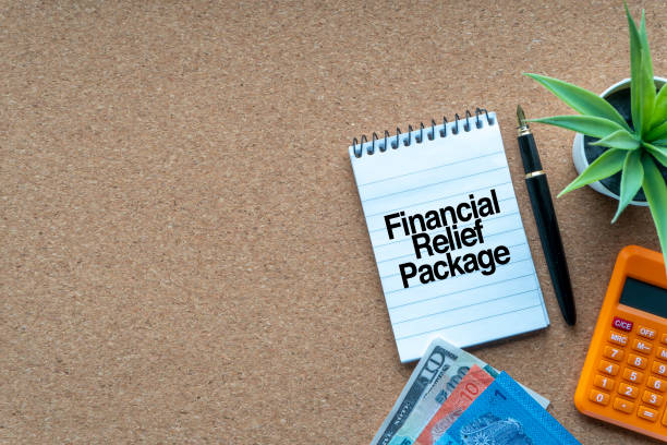 istock FINANCIAL RELIEF PACKAGE text with notepad, calculator, decorative plant, money banknotes, keyboard and fountain pen on wooden background.und 1329326113