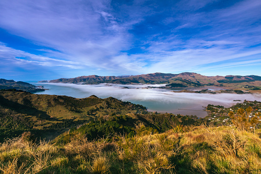This June 2021 image shows Te Whakaraupō Lyttelton Harbour, a part of Ōtautahi Christchurch, in Horomaka Banks Peninsula, Aotearoa New Zealand. Fog blankets part of the harbour. The Pacific Ocean is seen in the distance.