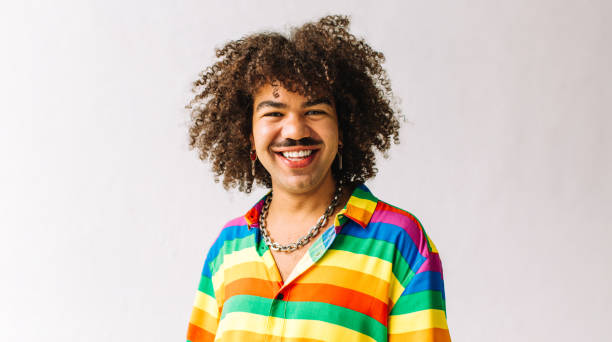 Gay man smiling in a studio Carefree young gay man celebrating gay pride in a studio. Young man smiling at the camera while standing alone against a white background. Young gay man wearing a shirt with rainbow colours. lgbtqia culture photos stock pictures, royalty-free photos & images