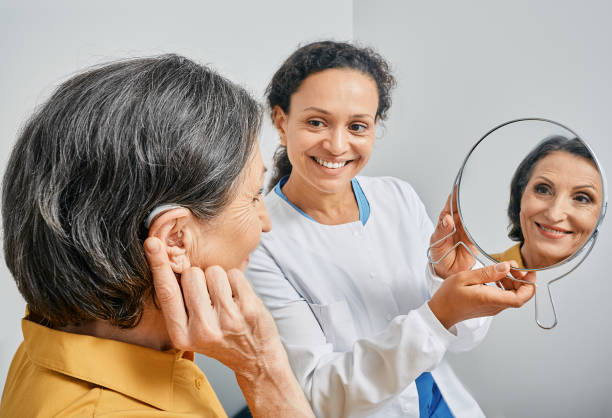 Mature woman with BTE hearing aid looks at himself in mirror held by smiling audiologist, and tries on hearing device. Stylish hearing device Mature woman with BTE hearing aid looks at himself in mirror held by smiling audiologist, and tries on hearing device. Stylish hearing device hearing aid photos stock pictures, royalty-free photos & images