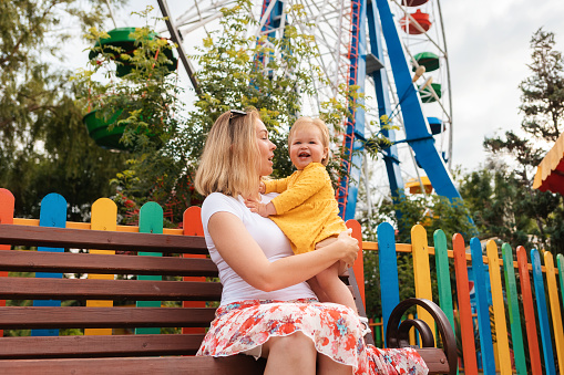 Caucasian young woman with a happy baby girl in her arms sitting on a bench. In the background, a colorful fence and ferris wheel. Concept of summer family vacation.
