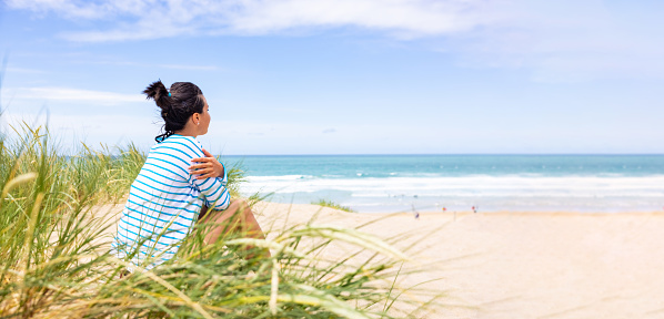 Woman at seaside in Cornwall looking at beach and waves - Rear side view of a woman sitting on sand dunes with ocean on background on a sunny day - Travel and happiness concepts