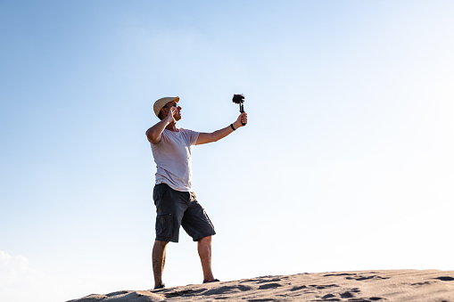Side view of male traveler standing on sand dune and filming content for social media