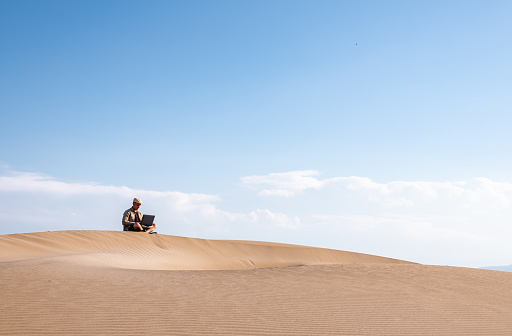 Front view of male traveler sitting on sandy dune using laptop