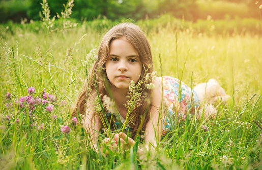 Girl with long brown hair is smiling while lying down on a lawn with outstretched arms and closed eyes with flowers in her hair,side view during summertime and daylight