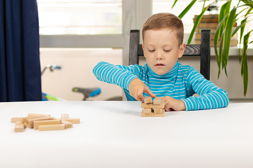 A cute seven-year-old child boy playing in wooden blocks  at home at a white wooden table against the background of a light window. Selective focus. Close-up. Portrait