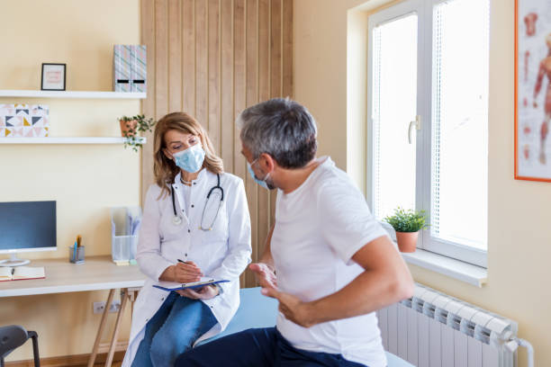 foto einer reifen ärztin mit einer schützenden gesichtsmaske, die anamnese von ihrer reifen patientin nimmt - doctors office examination room examination table office stock-fotos und bilder