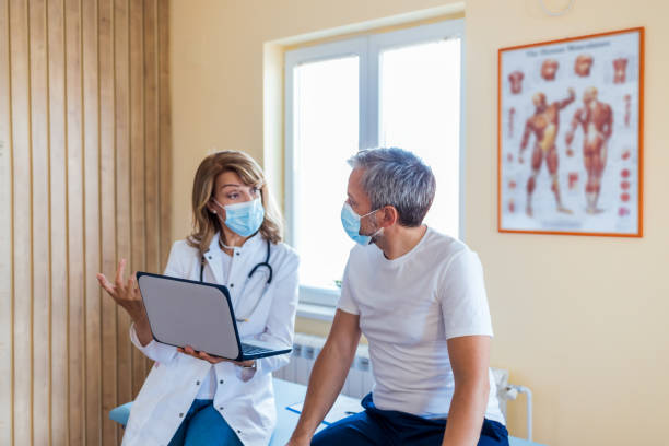 Photo of a female mature  doctor with a protective face mask taking anamnesis from her mature patient Mature Female doctor wearing face mask discussing with man patient at the hospital anamnesis stock pictures, royalty-free photos & images