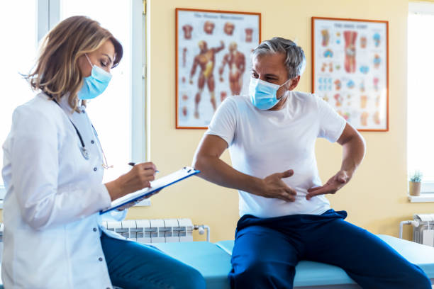 photo of a female mature  doctor with a protective face mask taking anamnesis from her mature patient - doctors office examination room examination table office imagens e fotografias de stock