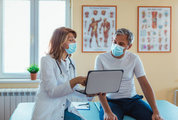 Photo of a female mature  doctor with a protective face mask taking anamnesis from her mature patient Mature Female doctor wearing face mask discussing with man patient at the hospital anamnesis stock pictures, royalty-free photos & images
