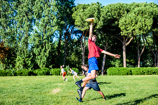 Group of happy friends playing flying disc in the park in summer time - Boy jump for take a frisbie - Sport, education, fun, team, lifestyle concept about healthy life in the outdoor