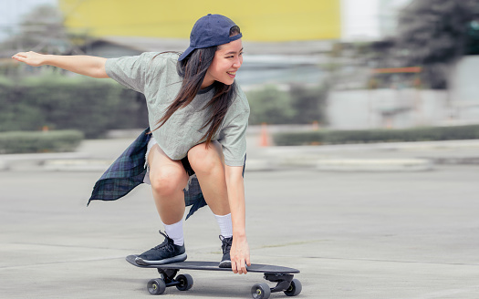 Beautiful happy Asian healthy woman smiling, motion speed riding and playing extreme sportive skateboard as outdoor activity with happiness, relaxation and fun during holidays in summer vacation