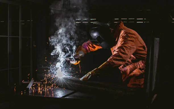 Photo of Asian handsome beard male mechanic wearing orange uniform and safety mask, using equipment and welding irons or metals with intention in industry factory.