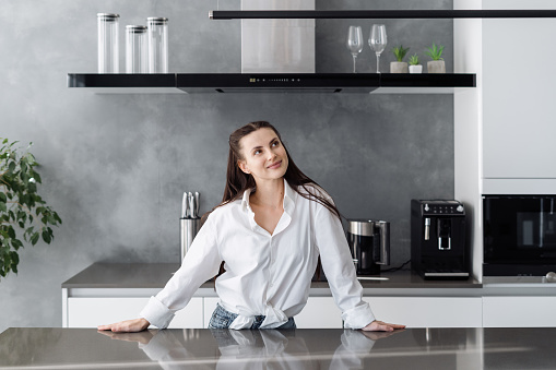 Young beautiful dreamy woman in casual outfit standing at table in modern fully furnished kitchen and thinking what to cook on breakfast for whole family on weekend