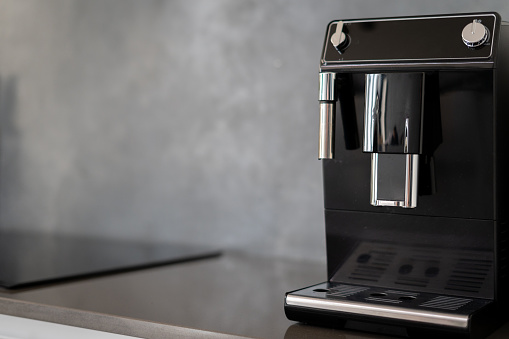 Black coffee making machine with glossy metal parts on shiny kitchen counter next to induction stovetop, pair of control knobs to change modes, blurred background, copy space for text