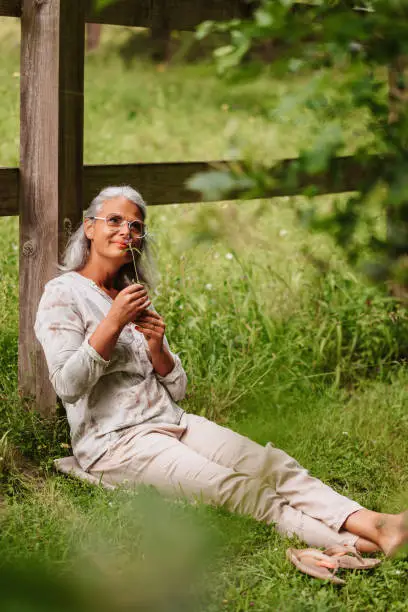 Spiritual mature woman with white hair sitting respectfully in the meadow with a wild flower in her hands appreciating gratefully the green nature and the life. Useful for mindfulness, conscious living and happy life style concepts. Creative color editing with slightly grain. Part of a series.
