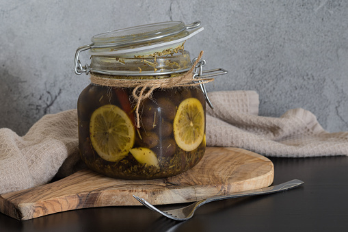Olives in a glass jar in oil with lemon slices on grey background.