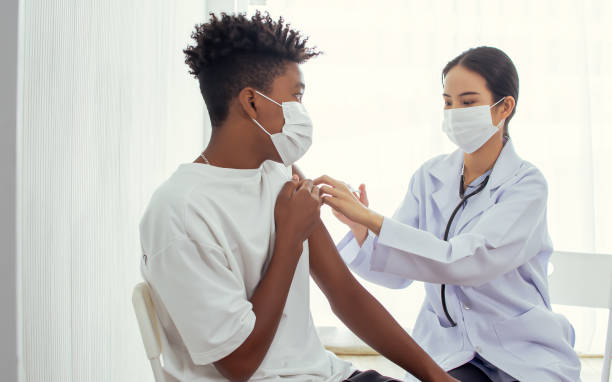 afrikanischer teenager, der eine gesichtsmaske trägt, sich impfen lassen, um viren zu schützen oder zu verhindern, angst vor der injektion von impfstoffen wird, während eine asiatische ärztin eine spritze vorbereitet, um den patienten im krankenhaus zu - grippeimpfstoff stock-fotos und bilder