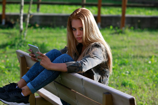 Serious young woman staring intently at the camera as she relaxes with her legs up over the back of a rustic wooden bench outdoors at sunset in a lush garden