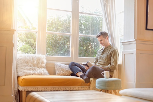 An attractive caucasian male seated in a reading nook. He is sitting with his legs crossed and reading a new book. Modern lounge room setting with the sun setting through the window.