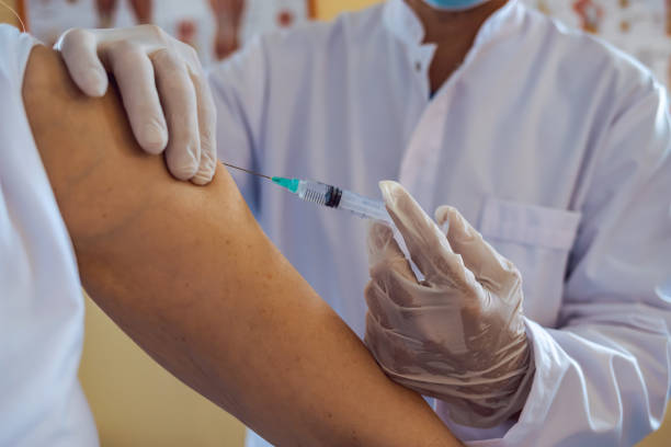 istock Close up of general practitioner hand holding vaccine injection while wearing face protective mask during covid-19 pandemic. Mature man nurse with surgical mask giving injection to senior woman. 1329394719