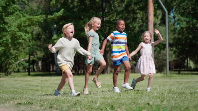 Tracking side view shot of group of happy multiethnic kids running on green grass holding hands. Diverse children hopping on lawn in park or woodland in slow motion and screaming with excitement