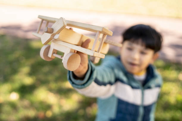 A close up of a wooden airplane being held high in the air by a young boy, the boy is out of focus, outside in a local park on a bright sunny day