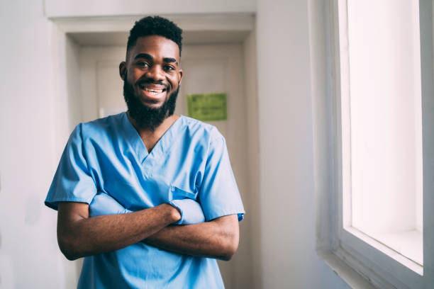 Smiling doctor standing with his arms crossed wearing protective gloves Smiling doctor standing with his arms crossed wearing protective gloves and hospital scrubs in a hospital corridor community outreach photos stock pictures, royalty-free photos & images