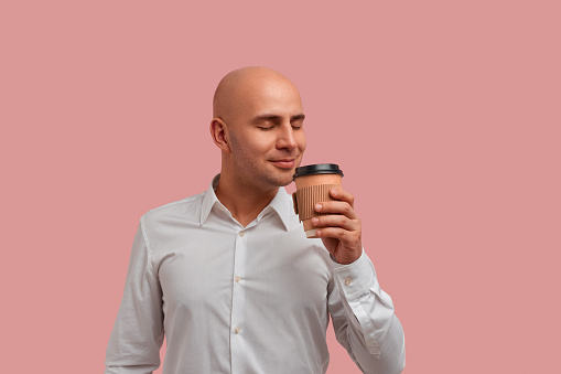 Enjoy drinking caffeine beverage. Attractive bald man with bristle, smells aromatic coffee, has break, closes eyes with pleasure, wears white shirt, isolated on pink background