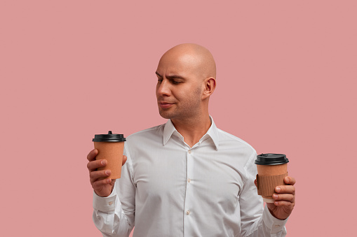What to choose. Thoughtful bald man with bristle, holds two paper cups with beverages, trying to select one. Tea or coffee. Wears white shirt, isolated on pink background