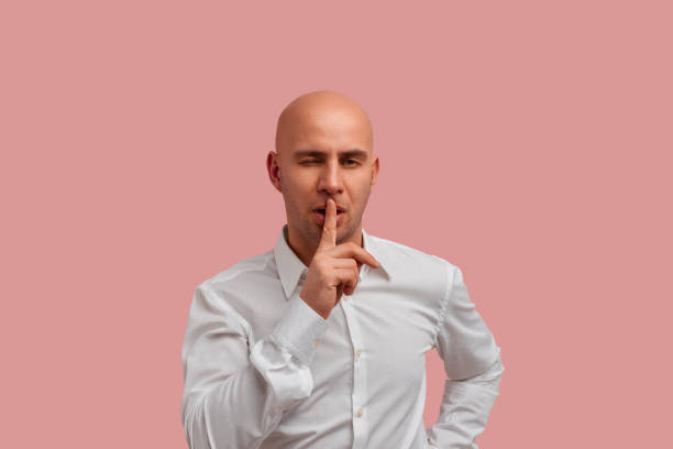 istock Do not tell anyone. Sexual winking looking bald man posing over pink studio background, makes hush gesture, looks at the camera, flirts with someone. Has bristle, dressed in white shirt. 1329401885